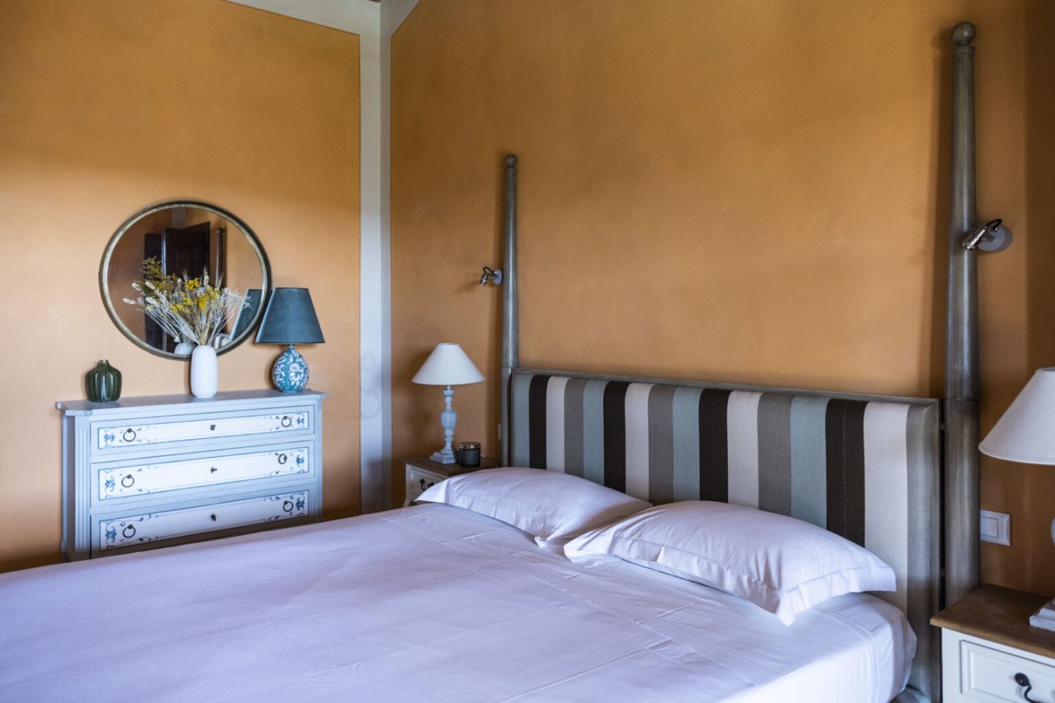 Cozy bedroom featuring a striped headboard, soft bedding, and an orange wall. The decor includes a round mirror, a blue and white vase, and stylish lamps on bedside tables, creating a warm and inviting atmosphere. | Only on SopranoVillas