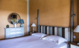 Cozy bedroom featuring a striped headboard, soft bedding, and an orange wall. The decor includes a round mirror, a blue and white vase, and stylish lamps on bedside tables, creating a warm and inviting atmosphere.