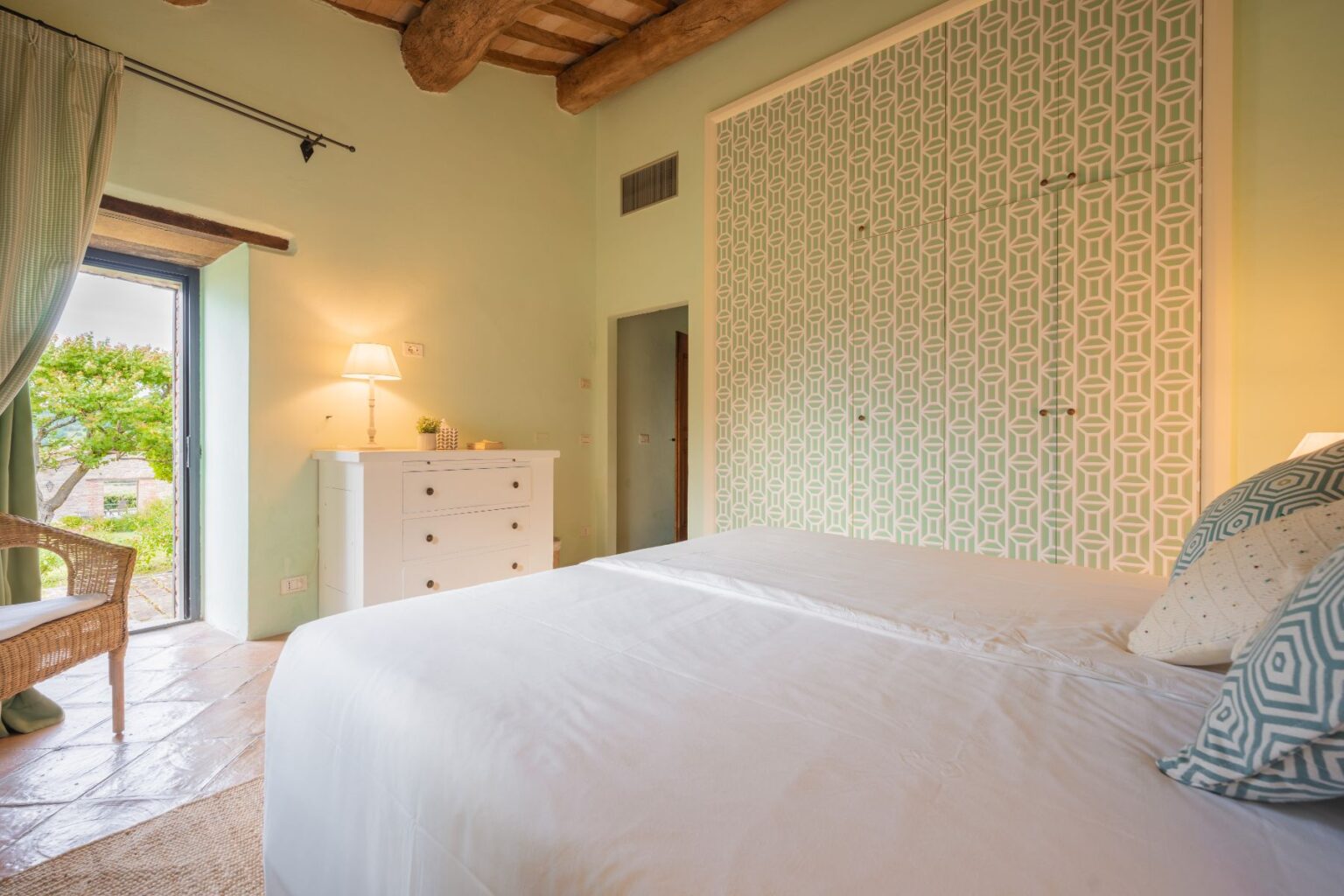 Cozy bedroom interior featuring a comfortable bed with white linens, decorative pillows, and a stylish patterned accent wall. Natural light streams in through an open door leading to a garden view, complemented by a white dresser and a lamp for a warm ambiance. Ideal for relaxation and tranquility. | Only on SopranoVillas