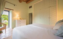 Cozy bedroom interior featuring a comfortable bed with white linens, decorative pillows, and a stylish patterned accent wall. Natural light streams in through an open door leading to a garden view, complemented by a white dresser and a lamp for a warm ambiance. Ideal for relaxation and tranquility.