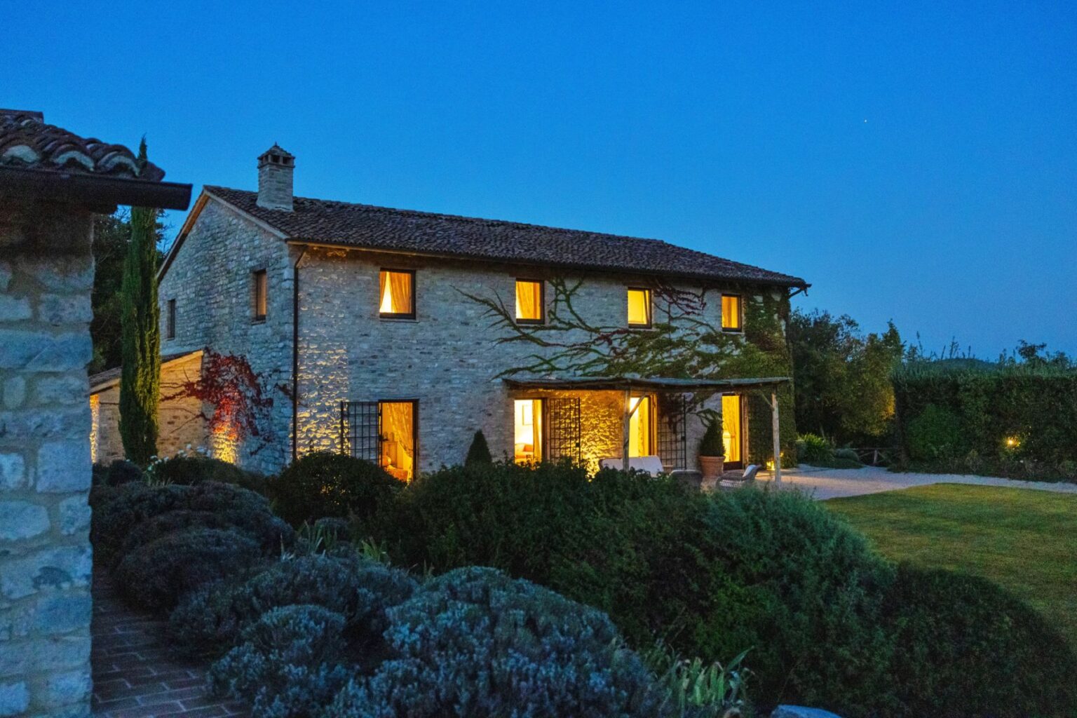 Stone farmhouse illuminated at dusk, featuring a rustic design with warm lights glowing from the windows, surrounded by lush greenery and well-maintained landscaping. | Only on SopranoVillas