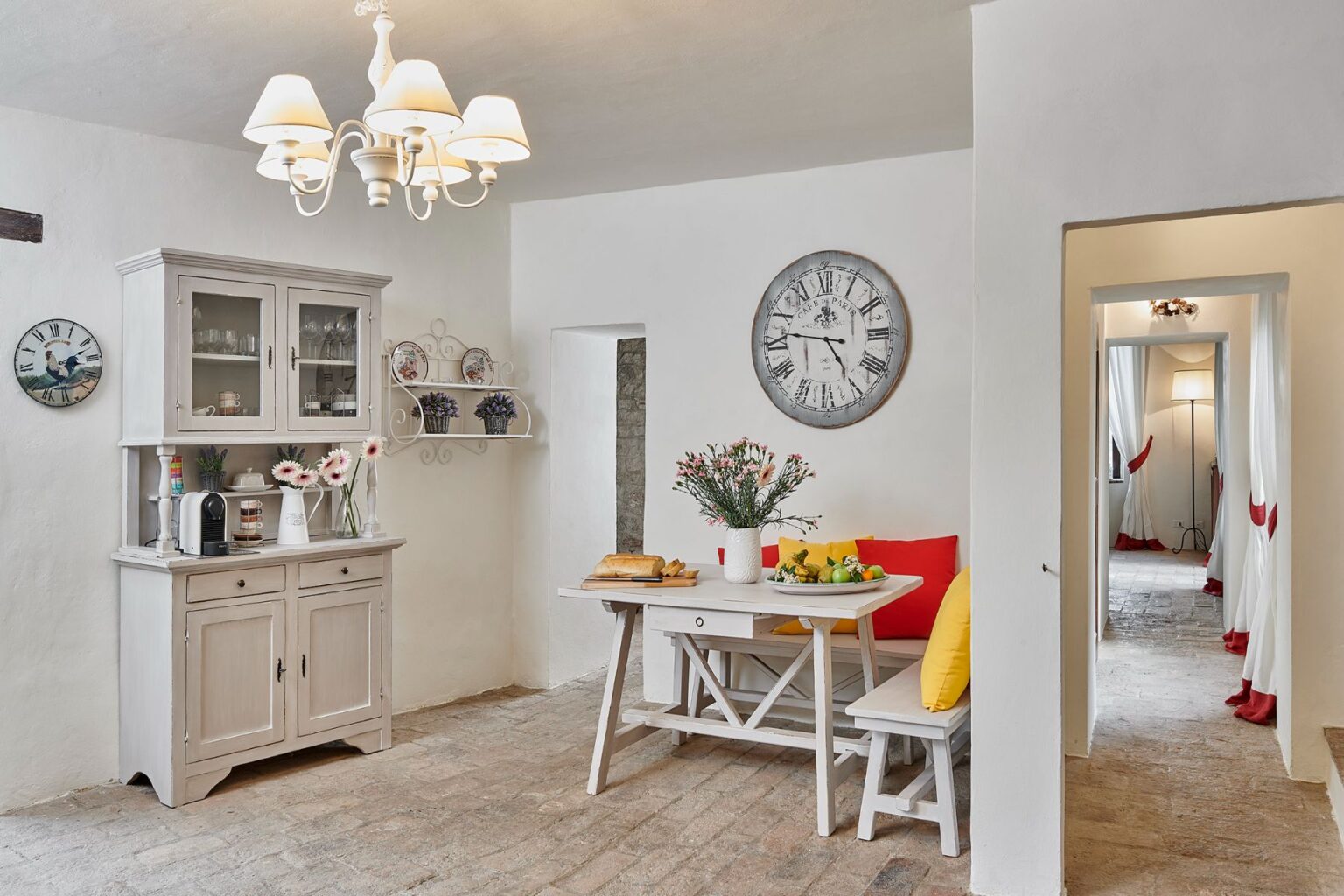 Bright and inviting kitchen interior featuring a wooden dining table with colorful cushions, a floral centerpiece, and a variety of fresh fruits. A light-colored cabinet displays glassware, while a vintage clock adorns the wall. The space is illuminated by a stylish chandelier, creating a warm atmosphere. | Only on SopranoVillas