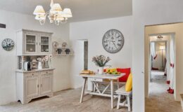 Bright and inviting kitchen interior featuring a wooden dining table with colorful cushions, a floral centerpiece, and a variety of fresh fruits. A light-colored cabinet displays glassware, while a vintage clock adorns the wall. The space is illuminated by a stylish chandelier, creating a warm atmosphere.