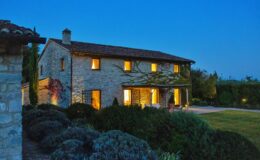 Stone farmhouse illuminated at dusk, featuring a rustic design with warm lights glowing from the windows, surrounded by lush greenery and well-maintained landscaping.