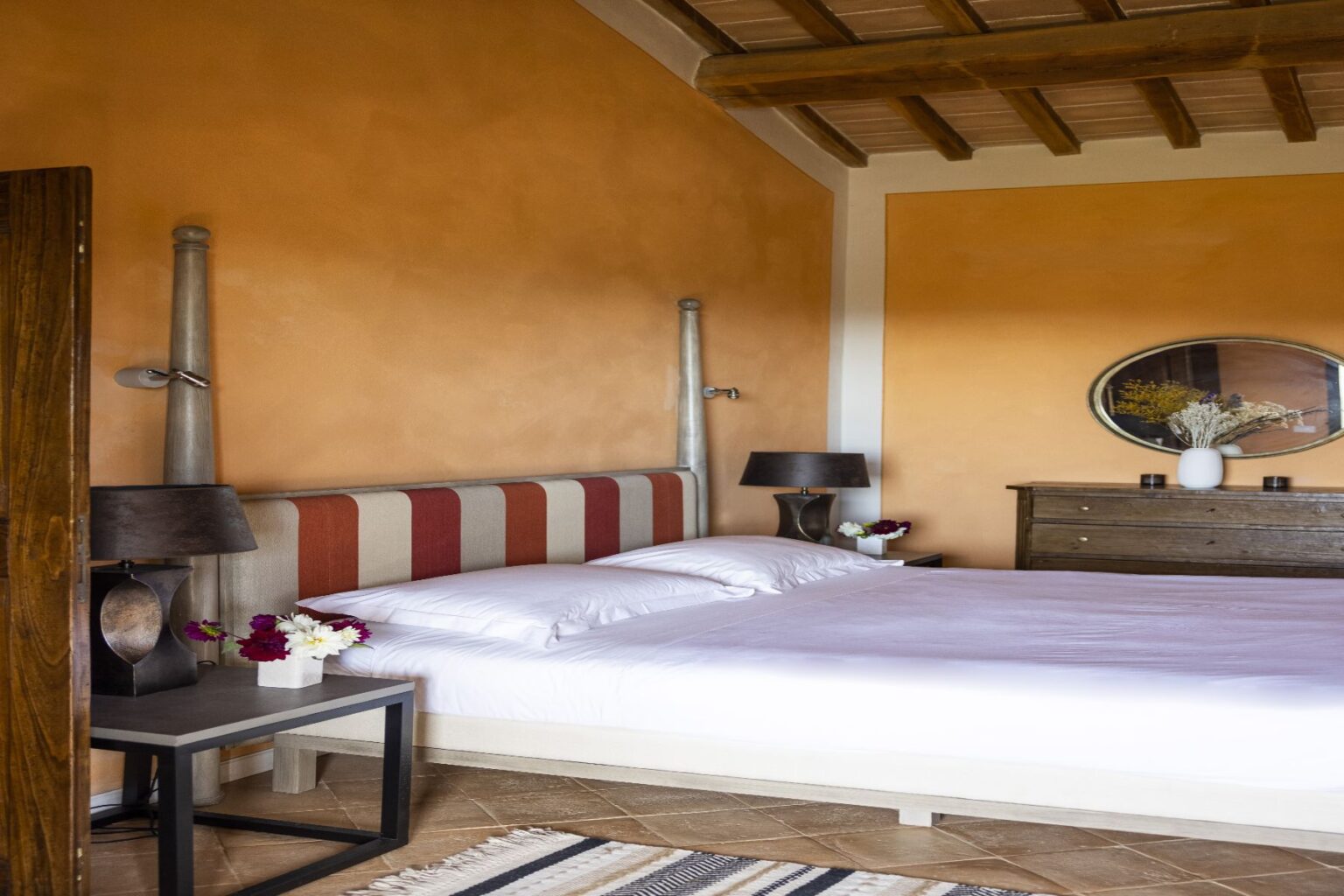 Cozy bedroom with a stylish striped headboard, white bedding, and decorative lamps on nightstands. The warm orange walls and wooden ceiling create a welcoming atmosphere, complemented by a vintage dresser and a round mirror with floral decor. | Only on SopranoVillas