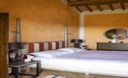 Cozy bedroom with a stylish striped headboard, white bedding, and decorative lamps on nightstands. The warm orange walls and wooden ceiling create a welcoming atmosphere, complemented by a vintage dresser and a round mirror with floral decor.