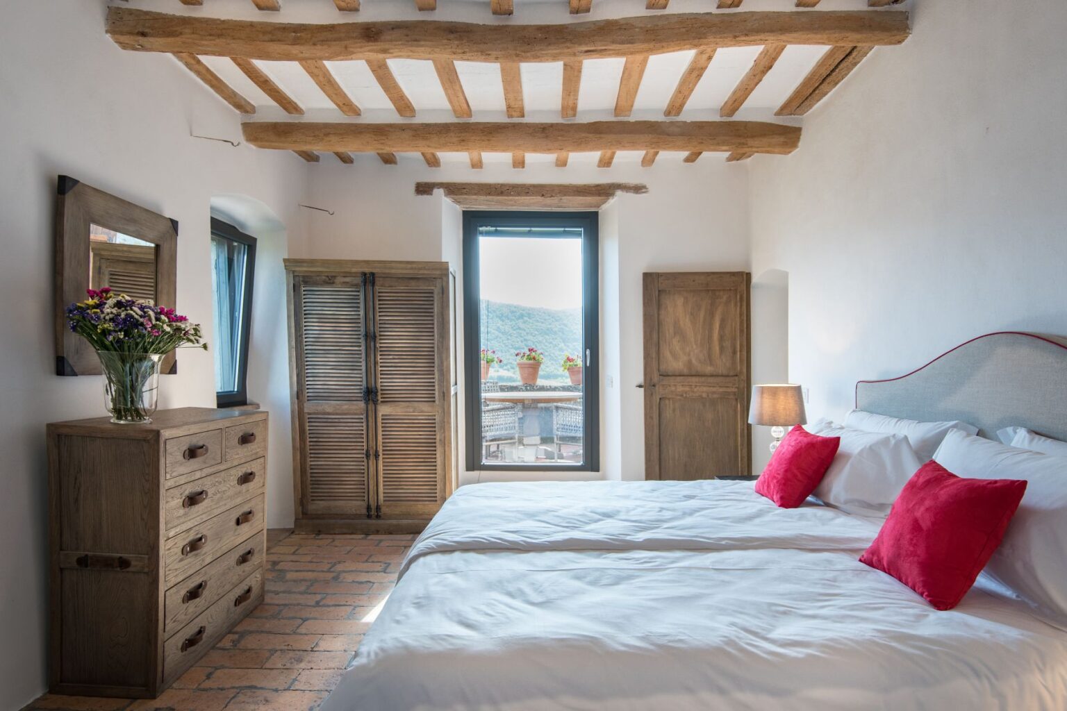 Cozy bedroom featuring two twin beds with white linens and red accent pillows, a wooden dresser, and a wardrobe. Natural light streams through a large window, highlighting rustic wooden beams and terracotta flooring. A vase of colorful flowers adds a touch of decor. Ideal for a relaxing getaway. | Only on SopranoVillas