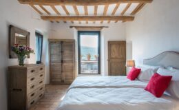 Cozy bedroom featuring two twin beds with white linens and red accent pillows, a wooden dresser, and a wardrobe. Natural light streams through a large window, highlighting rustic wooden beams and terracotta flooring. A vase of colorful flowers adds a touch of decor. Ideal for a relaxing getaway.