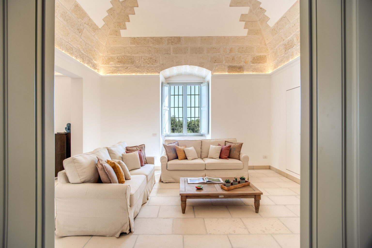Spacious living room with a modern design featuring a comfortable white sofa, decorative cushions, and a wooden coffee table, illuminated by natural light from a large window. Stone walls and elegant ceiling details enhance the inviting atmosphere. | Only on SopranoVillas