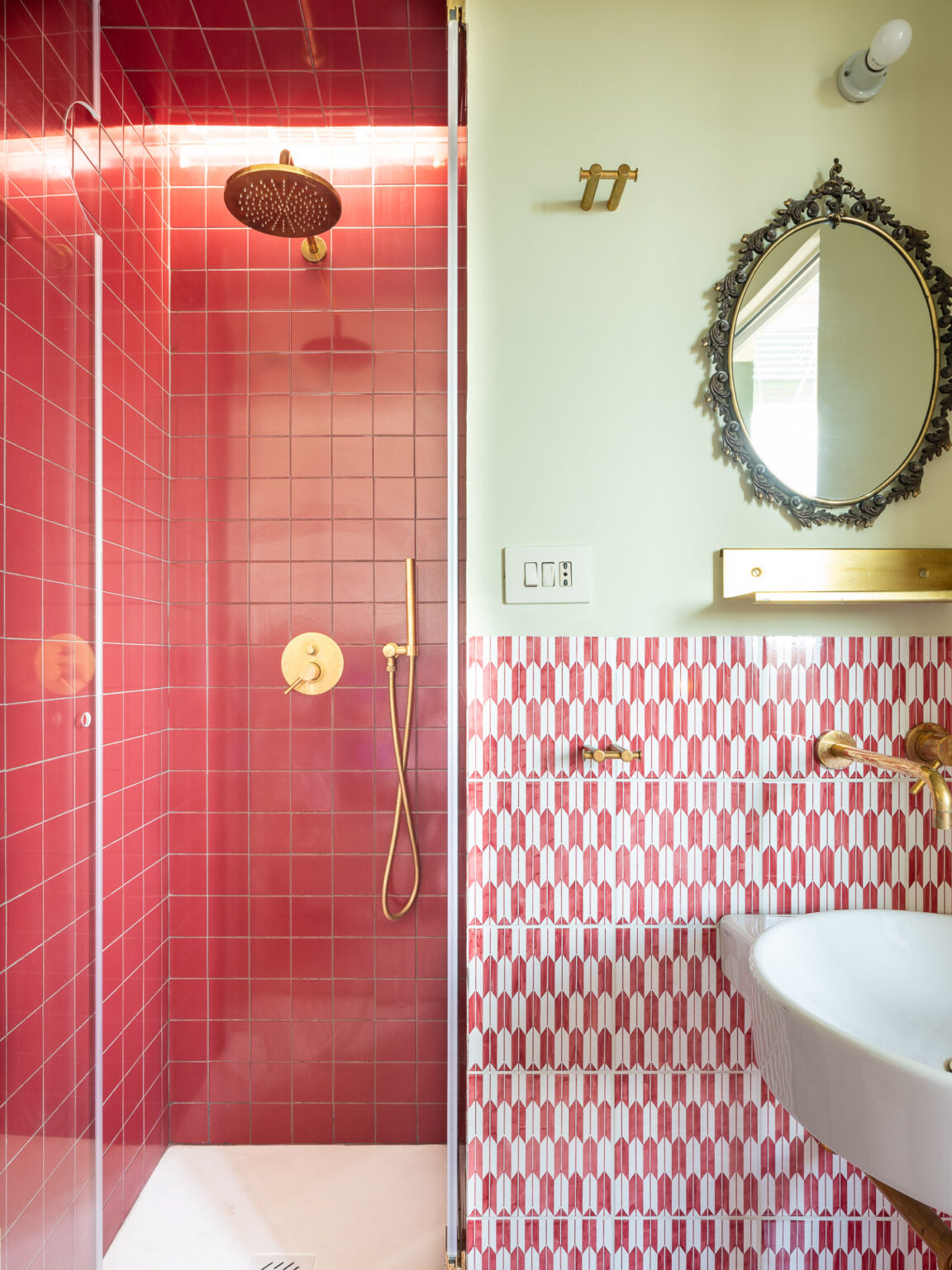 Modern bathroom design featuring a vibrant red tiled shower with a rain showerhead, complemented by a stylish gold faucet. The adjacent wall showcases a unique patterned tile design in shades of red, while a decorative mirror adds elegance to the space. Soft green walls enhance the overall aesthetic, creating a chic and contemporary atmosphere. | Only on SopranoVillas