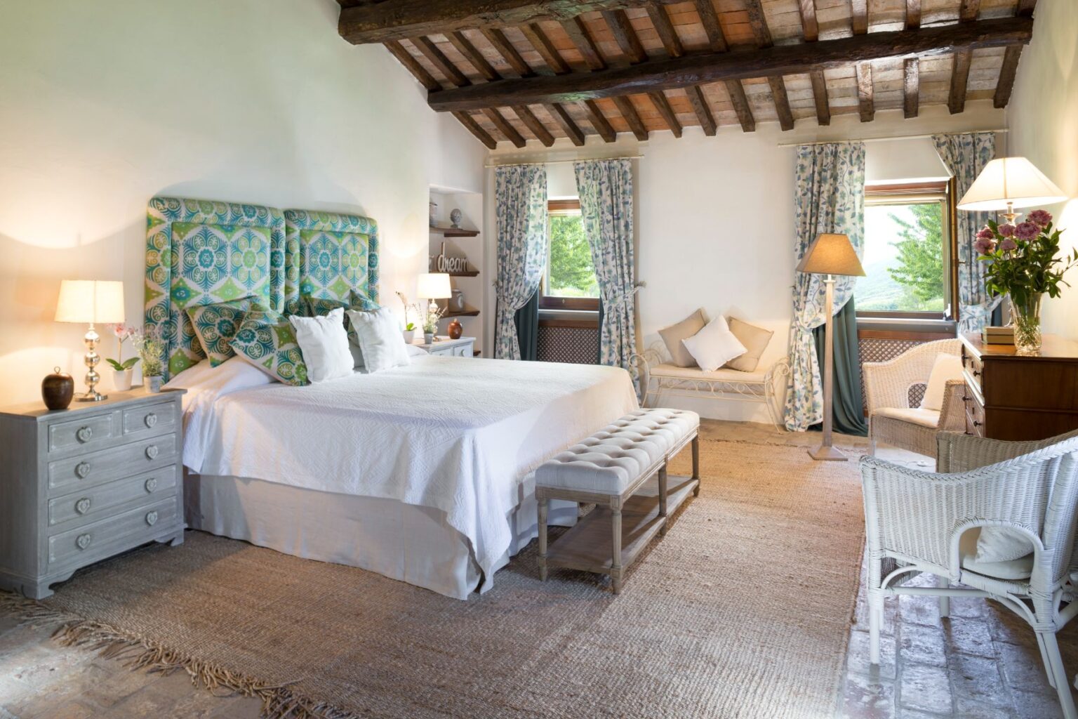 Bright and airy bedroom featuring a large bed with a colorful patterned headboard, complemented by white linens and decorative pillows. The room includes a stylish wooden dresser, a cozy seating area with a bench and wicker chairs, and elegant curtains. Natural light pours in through the windows, enhancing the inviting atmosphere. Perfect for a relaxing getaway. | Only on SopranoVillas