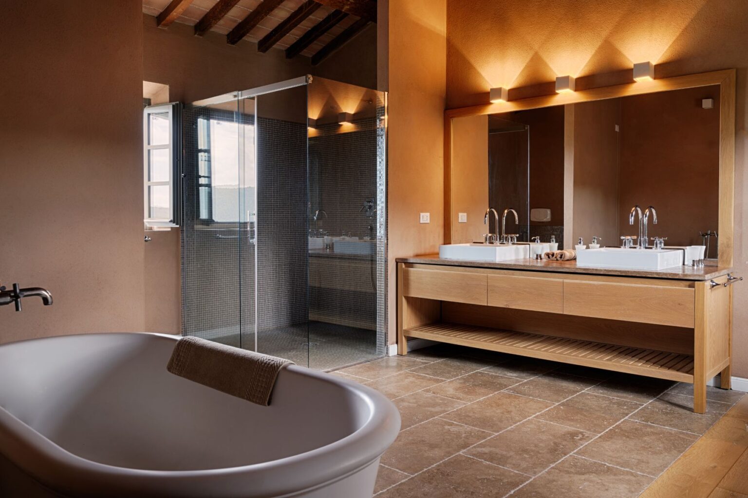 Modern bathroom featuring a freestanding bathtub, double sink vanity with elegant fixtures, and a glass shower enclosure. The warm, earthy tones and natural materials create a serene and stylish atmosphere, complemented by natural light from the window. | Only on SopranoVillas