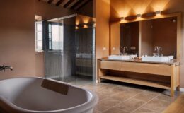 Modern bathroom featuring a freestanding bathtub, double sink vanity with elegant fixtures, and a glass shower enclosure. The warm, earthy tones and natural materials create a serene and stylish atmosphere, complemented by natural light from the window.