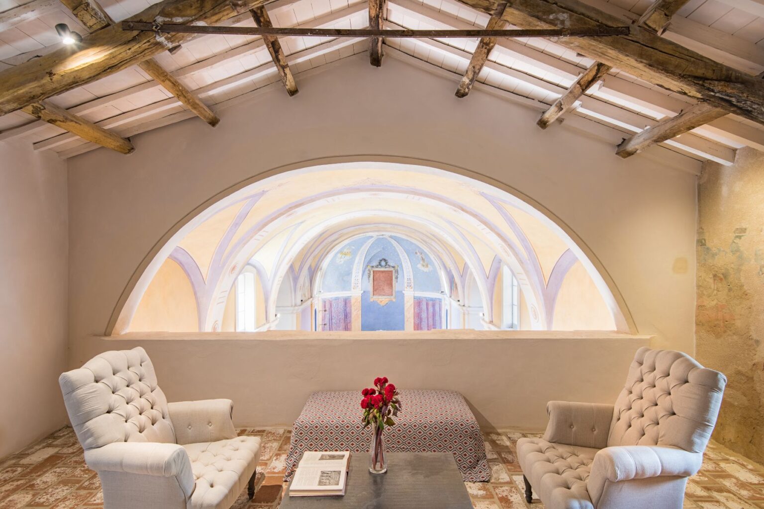 Cozy interior of a stylish loft featuring two upholstered chairs, a decorative table with a floral arrangement, and a stunning arched window showcasing colorful frescoes and architectural details in the background. | Only on SopranoVillas