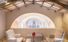 Cozy interior of a stylish loft featuring two upholstered chairs, a decorative table with a floral arrangement, and a stunning arched window showcasing colorful frescoes and architectural details in the background.