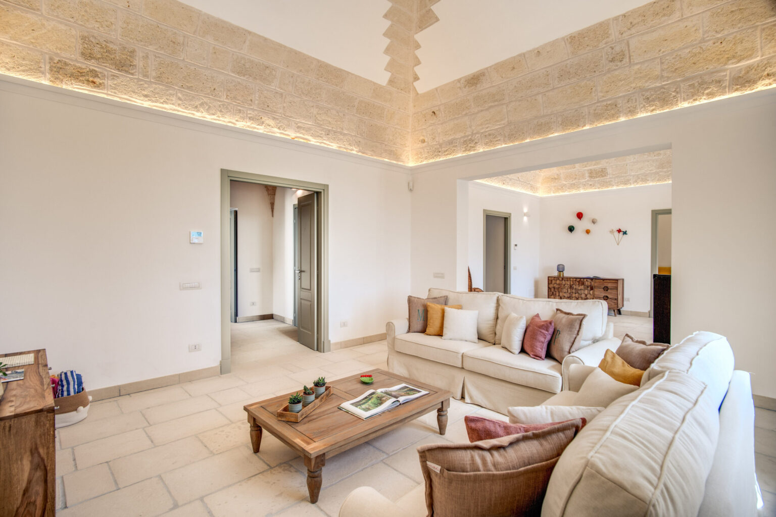 Modern living room featuring a comfortable beige sofa with colorful cushions, a wooden coffee table, and natural stone walls. The space is illuminated by soft lighting, creating a warm and inviting atmosphere, with doorways leading to other rooms. | Only on SopranoVillas