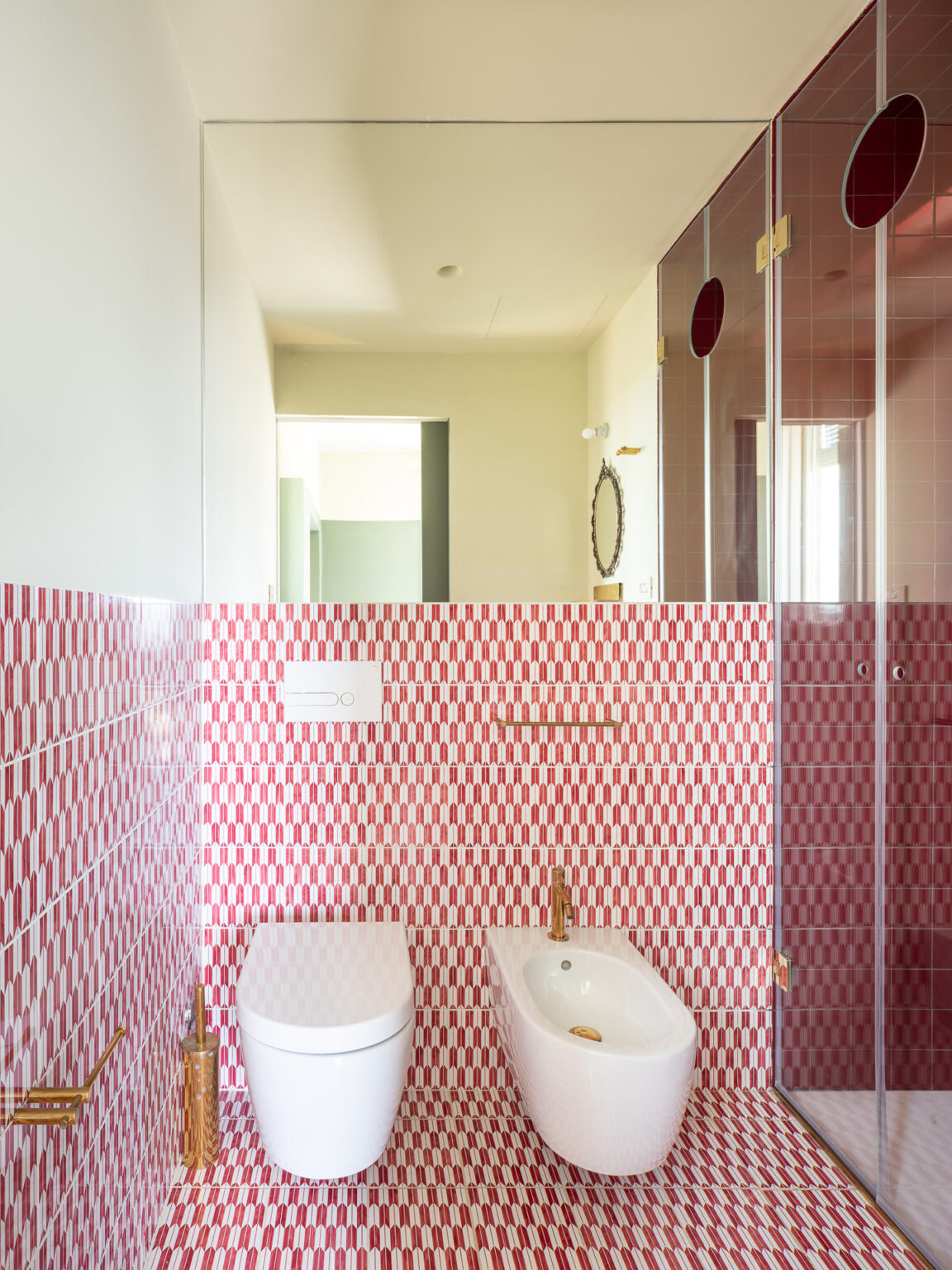 Modern bathroom design featuring a bidet and toilet with elegant gold fixtures, surrounded by vibrant red and white patterned tiles. A large mirror reflects the spacious layout, complemented by a glass shower enclosure. | Only on SopranoVillas
