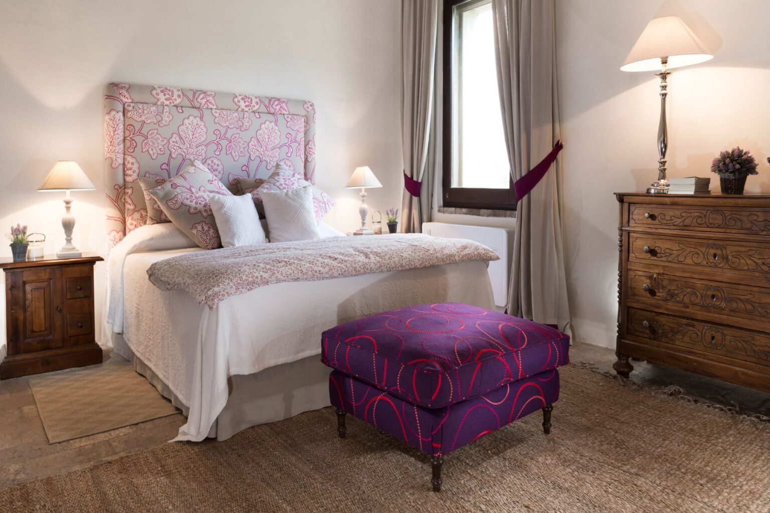 Cozy bedroom interior featuring a floral upholstered headboard, white bedding, and decorative pillows. A wooden nightstand with a lamp and a small plant complements the space, while a vibrant purple ottoman adds a pop of color. Natural light streams through a window, enhancing the warm ambiance. | Only on SopranoVillas
