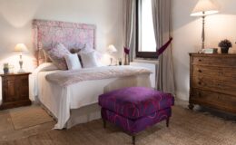 Cozy bedroom interior featuring a floral upholstered headboard, white bedding, and decorative pillows. A wooden nightstand with a lamp and a small plant complements the space, while a vibrant purple ottoman adds a pop of color. Natural light streams through a window, enhancing the warm ambiance.