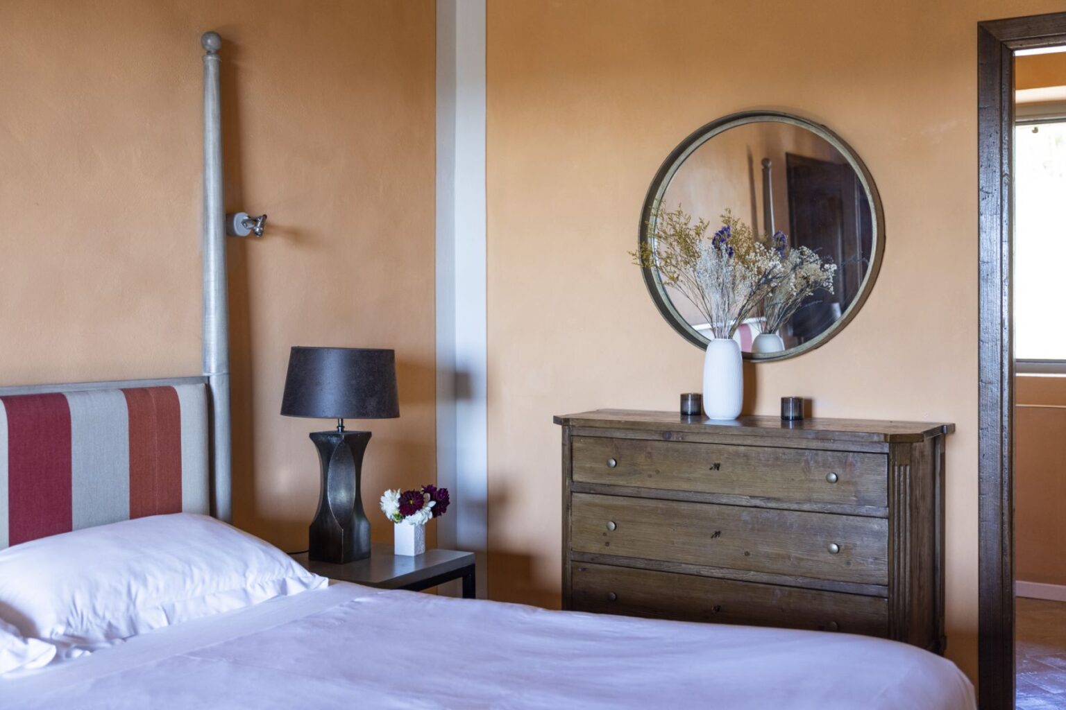 Cozy bedroom featuring a striped headboard, a wooden dresser, a round mirror, and decorative flowers. The warm orange walls create an inviting atmosphere, complemented by a stylish bedside lamp. | Only on SopranoVillas