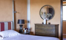 Cozy bedroom featuring a striped headboard, a wooden dresser, a round mirror, and decorative flowers. The warm orange walls create an inviting atmosphere, complemented by a stylish bedside lamp.