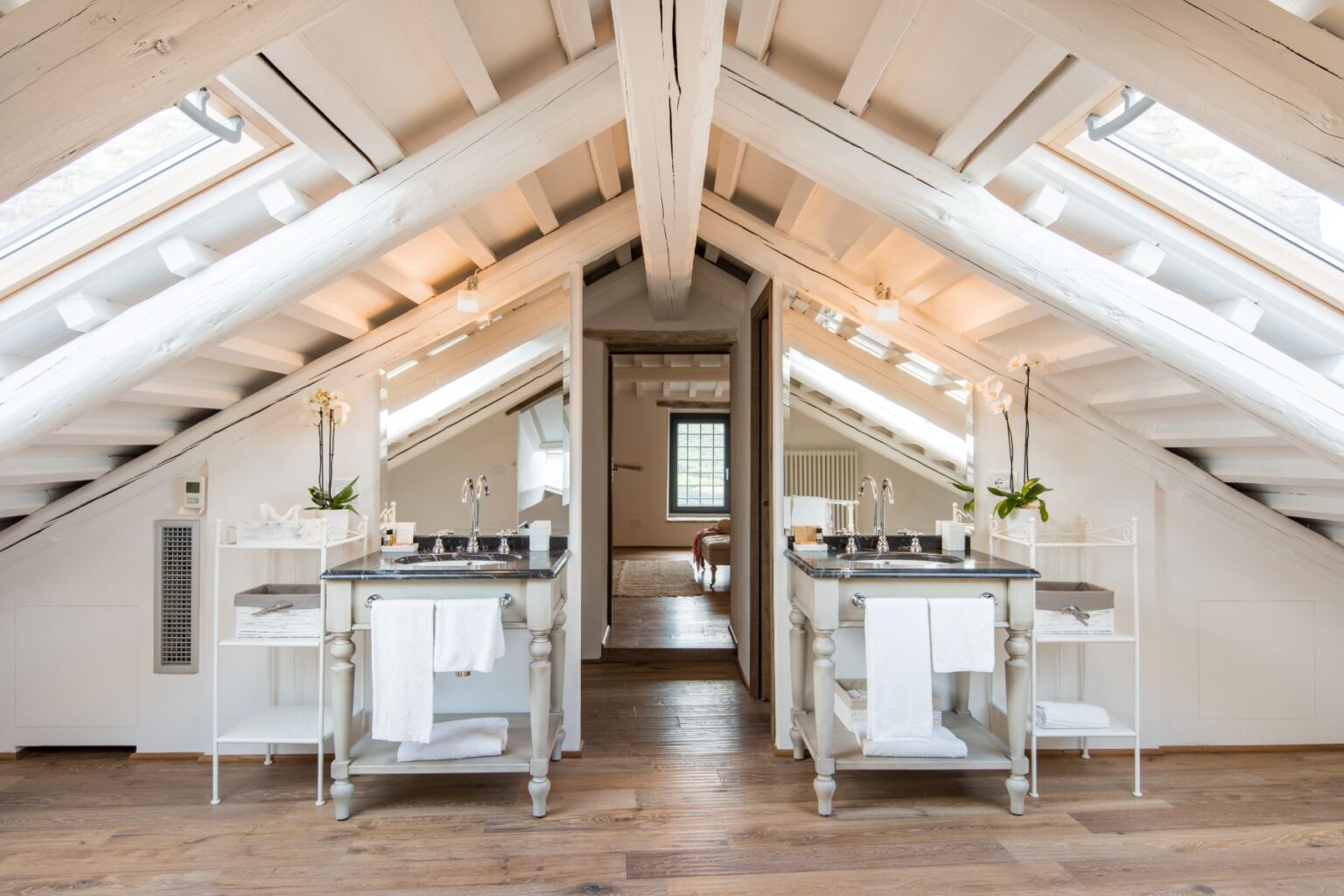 Bright and modern attic bathroom featuring dual vanities with elegant fixtures, large mirrors, and natural light from skylights. The space showcases wooden beams, warm wood flooring, and minimalist decor, creating a serene and stylish atmosphere. | Only on SopranoVillas