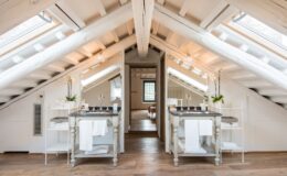 Bright and modern attic bathroom featuring dual vanities with elegant fixtures, large mirrors, and natural light from skylights. The space showcases wooden beams, warm wood flooring, and minimalist decor, creating a serene and stylish atmosphere.