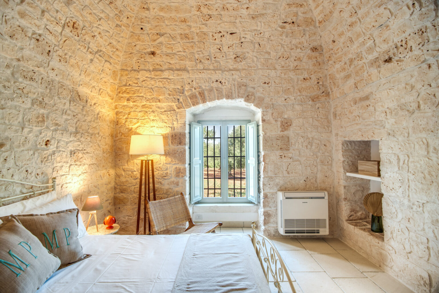Cozy bedroom featuring exposed stone walls, a decorative bed with embroidered pillows, a stylish floor lamp, and a large window with green shutters providing natural light and a view of the outdoors. | Only on SopranoVillas