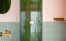 Modern bathroom featuring a green tiled shower with a gold showerhead, a white bathtub, and intricate green patterned tiles on the wall. Soft pink walls add warmth to the contemporary design.