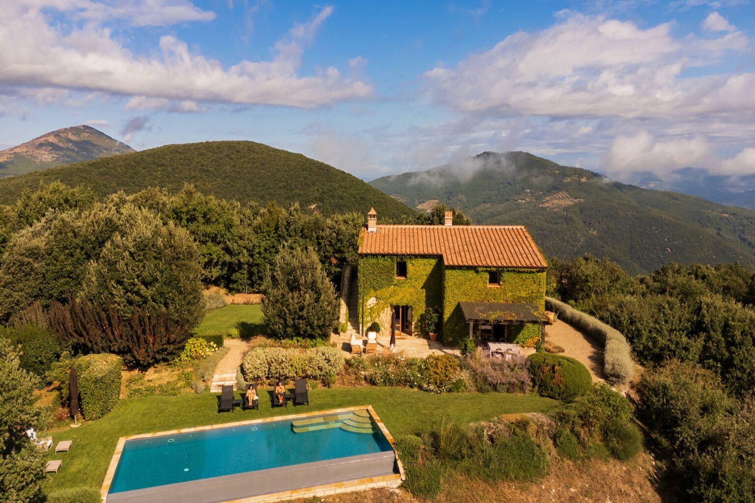 Aerial view of a charming ivy-covered villa with a swimming pool, surrounded by lush greenery and rolling hills in a picturesque landscape. Two people relax by the pool under the sun. Ideal for vacation rentals or countryside retreats. | Only on SopranoVillas