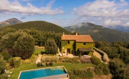 Aerial view of a charming ivy-covered villa with a swimming pool, surrounded by lush greenery and rolling hills in a picturesque landscape. Two people relax by the pool under the sun. Ideal for vacation rentals or countryside retreats.