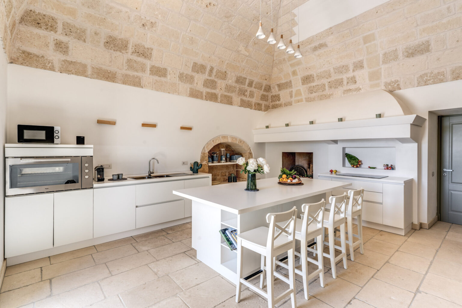 Modern kitchen interior featuring white cabinetry, a spacious island with seating, and stone walls. The design includes a microwave, sink, and decorative elements, creating a bright and inviting atmosphere. | Only on SopranoVillas