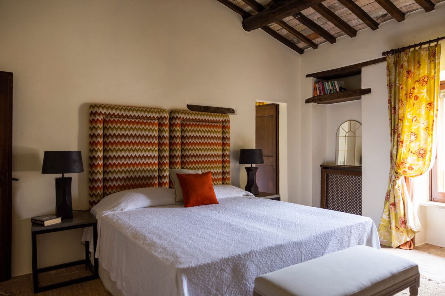 Cozy bedroom featuring a stylish bed with patterned headboards, a white quilt, and an accent pillow. The room includes a bedside lamp, a small bookshelf, and vibrant floral curtains that frame a window, creating a warm and inviting atmosphere. | Only on SopranoVillas