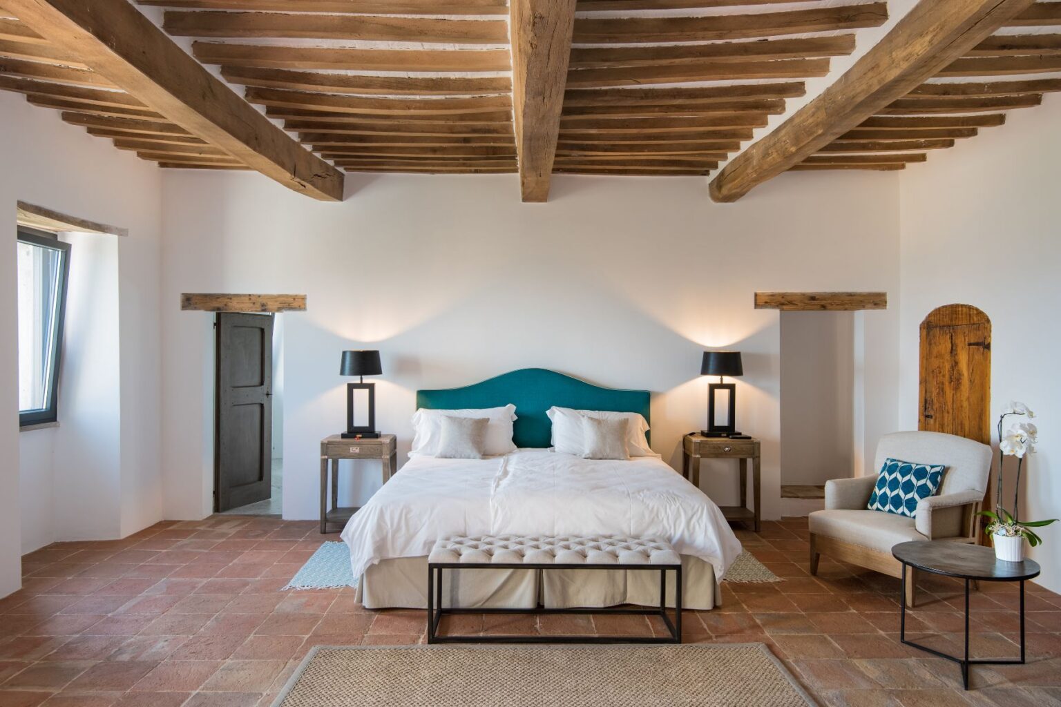 Spacious and modern bedroom featuring a king-size bed with a teal headboard, wooden beams, and warm lighting. The room includes bedside tables with lamps, a cozy armchair, and decorative elements like a rug and potted orchid, creating a relaxing atmosphere. | Only on SopranoVillas
