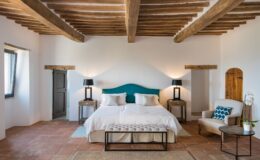 Spacious and modern bedroom featuring a king-size bed with a teal headboard, wooden beams, and warm lighting. The room includes bedside tables with lamps, a cozy armchair, and decorative elements like a rug and potted orchid, creating a relaxing atmosphere.