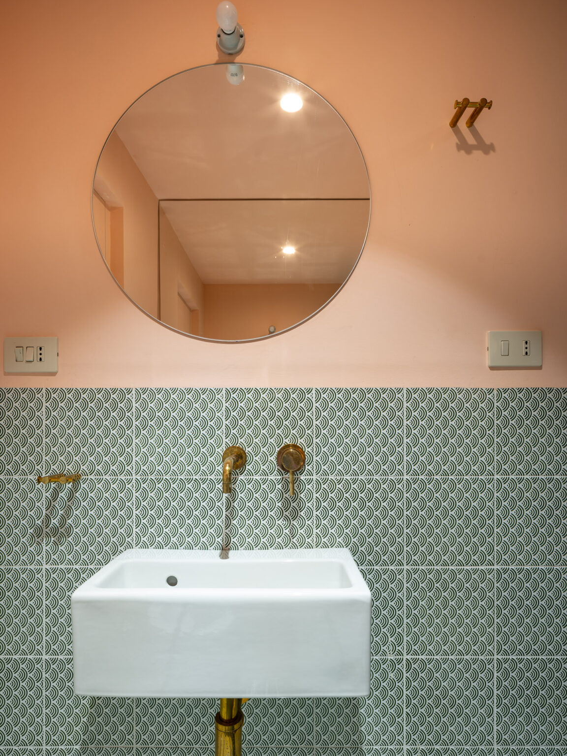 Modern bathroom with a round mirror, white sink, and decorative green patterned tiles. The warm peach wall complements the brass fixtures and lighting, creating a stylish and inviting space. | Only on SopranoVillas
