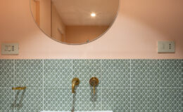 Modern bathroom with a round mirror, white sink, and decorative green patterned tiles. The warm peach wall complements the brass fixtures and lighting, creating a stylish and inviting space.