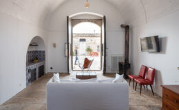 Interior view of a stylish living space featuring a white sofa, red chairs, and a vintage wooden dresser. The room has high arched ceilings and an open door leading to a courtyard with red outdoor furniture. A rustic kitchen area with decorative tile and a television mounted on the wall enhance the modern yet cozy atmosphere.