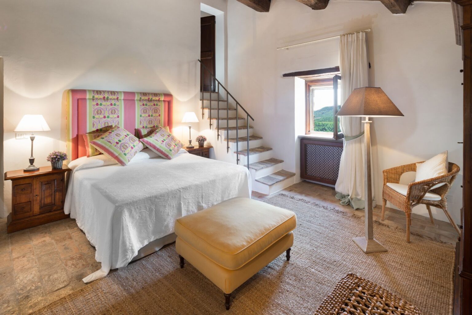 Cozy bedroom featuring a double bed with colorful patterned pillows, wooden nightstands with lamps, a comfortable ottoman, and a wicker chair. The room has a rustic staircase leading to an upper level and a window showcasing a scenic view of greenery. | Only on SopranoVillas