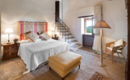 Cozy bedroom featuring a double bed with colorful patterned pillows, wooden nightstands with lamps, a comfortable ottoman, and a wicker chair. The room has a rustic staircase leading to an upper level and a window showcasing a scenic view of greenery.