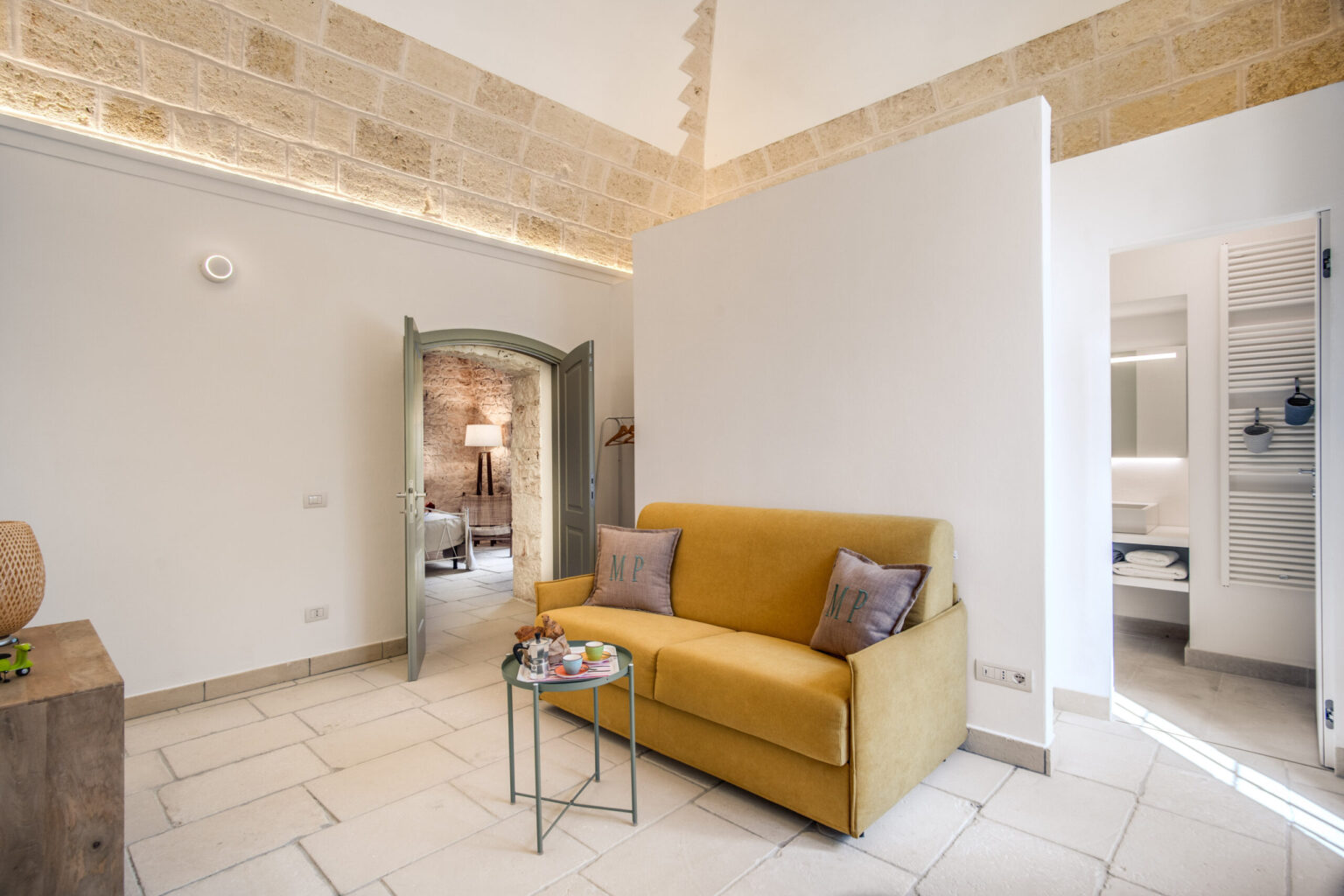 Cozy interior of a modern living space featuring a yellow sofa with decorative pillows, a small green side table with refreshments, and a doorway leading to a well-lit bedroom area with stone walls and stylish decor. The room has a bright and airy atmosphere, enhanced by natural light and contemporary design elements. | Only on SopranoVillas