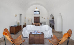 Bright and spacious living room featuring a white sofa with decorative pillows, two stylish leather chairs, a wooden coffee table with a magazine, and a wooden dining table in the background. The room showcases arched ceilings, white walls, and vintage furniture, providing a cozy and inviting atmosphere.