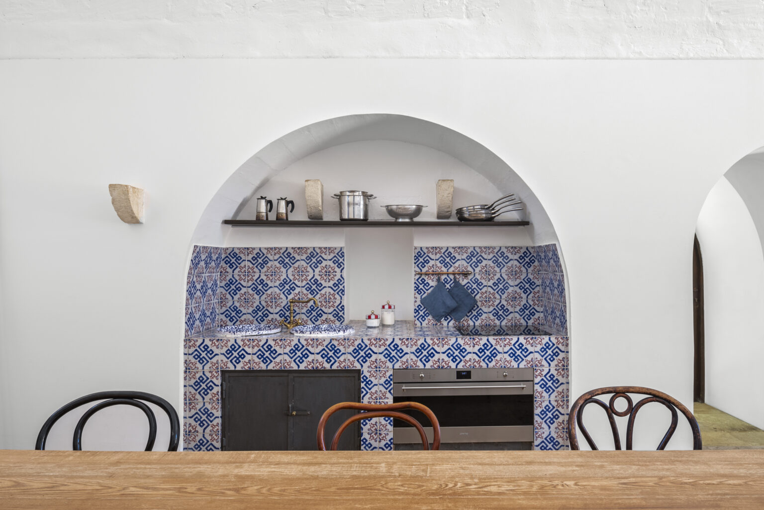 Modern kitchen interior featuring blue and white patterned tiles, a wooden dining table, and elegant archways. The kitchen is equipped with stainless steel cookware, a built-in oven, and minimalist decor, creating a stylish and inviting atmosphere. | Only on SopranoVillas