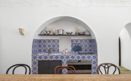 Modern kitchen interior featuring blue and white patterned tiles, a wooden dining table, and elegant archways. The kitchen is equipped with stainless steel cookware, a built-in oven, and minimalist decor, creating a stylish and inviting atmosphere.