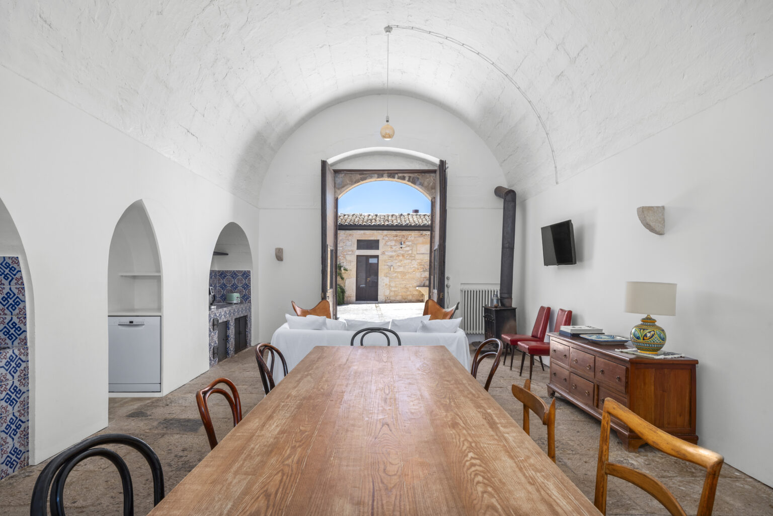 Spacious dining area with a wooden table and various chairs, featuring a rustic arched ceiling and large open doors leading to an outdoor courtyard. The room includes a kitchen area with decorative tiles, a modern refrigerator, and a cozy seating arrangement, creating a warm and inviting atmosphere. | Only on SopranoVillas