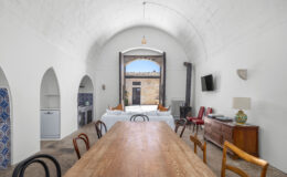 Spacious dining area with a wooden table and various chairs, featuring a rustic arched ceiling and large open doors leading to an outdoor courtyard. The room includes a kitchen area with decorative tiles, a modern refrigerator, and a cozy seating arrangement, creating a warm and inviting atmosphere.