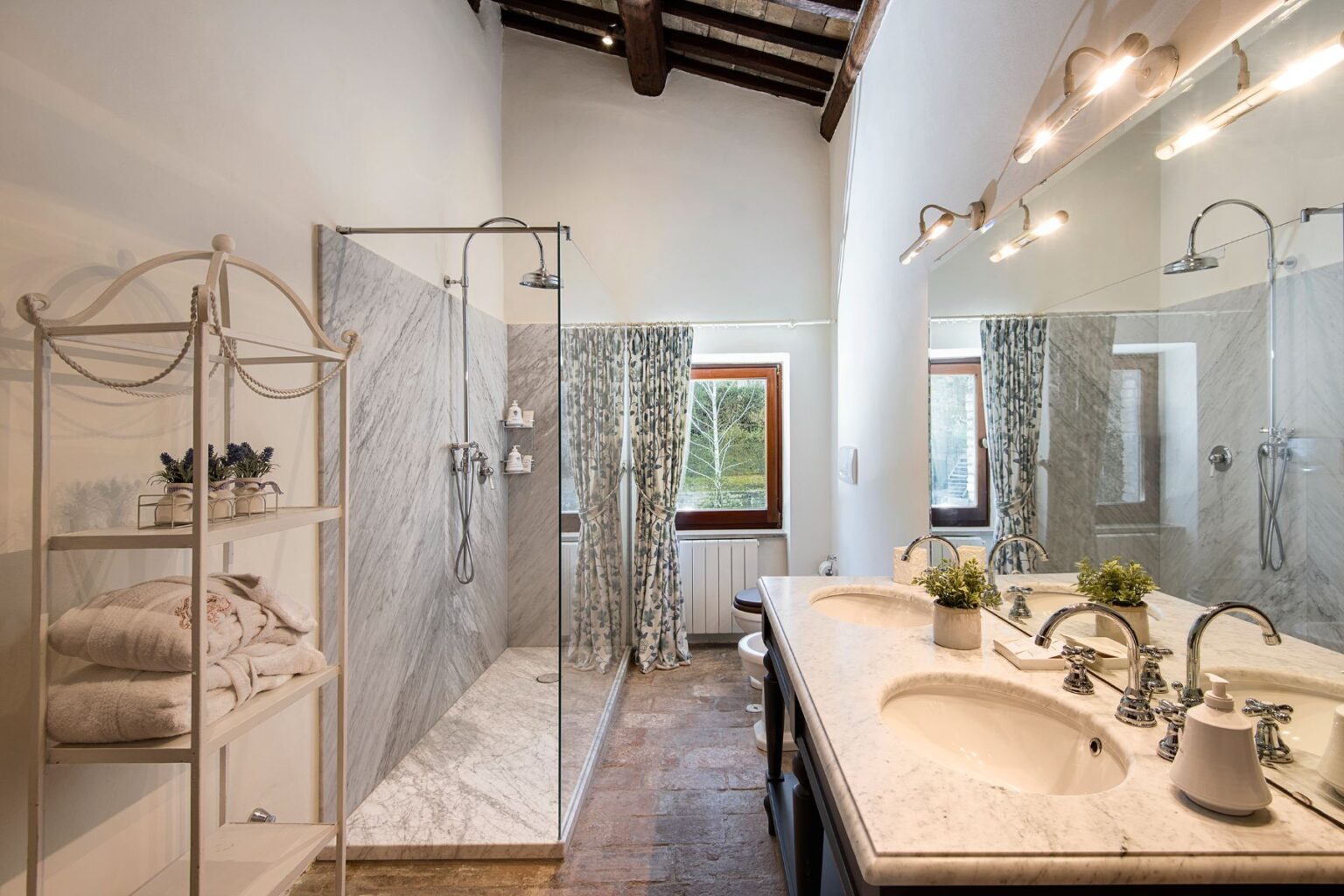 Modern bathroom featuring a spacious walk-in shower with marble walls, double sink vanity with elegant fixtures, and natural light from windows adorned with floral curtains. A stylish shelving unit displays neatly folded towels and decorative plants, enhancing the room's aesthetic. | Only on SopranoVillas