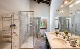 Modern bathroom featuring a spacious walk-in shower with marble walls, double sink vanity with elegant fixtures, and natural light from windows adorned with floral curtains. A stylish shelving unit displays neatly folded towels and decorative plants, enhancing the room's aesthetic.