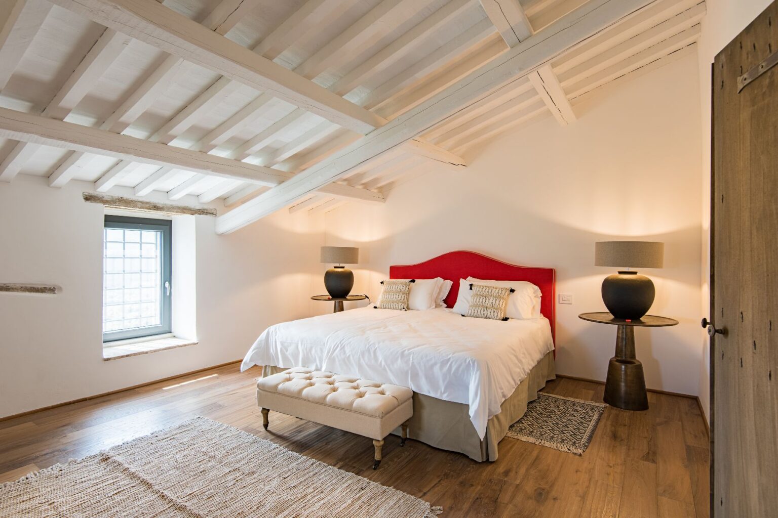 Modern bedroom interior featuring a plush king-size bed with a red headboard, stylish bedside lamps, and a cozy seating area. The room showcases wooden beams, a large window with a view, and warm wooden flooring, creating a serene and inviting atmosphere. Ideal for relaxation and comfort. | Only on SopranoVillas