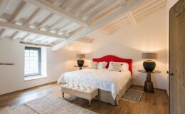 Modern bedroom interior featuring a plush king-size bed with a red headboard, stylish bedside lamps, and a cozy seating area. The room showcases wooden beams, a large window with a view, and warm wooden flooring, creating a serene and inviting atmosphere. Ideal for relaxation and comfort.